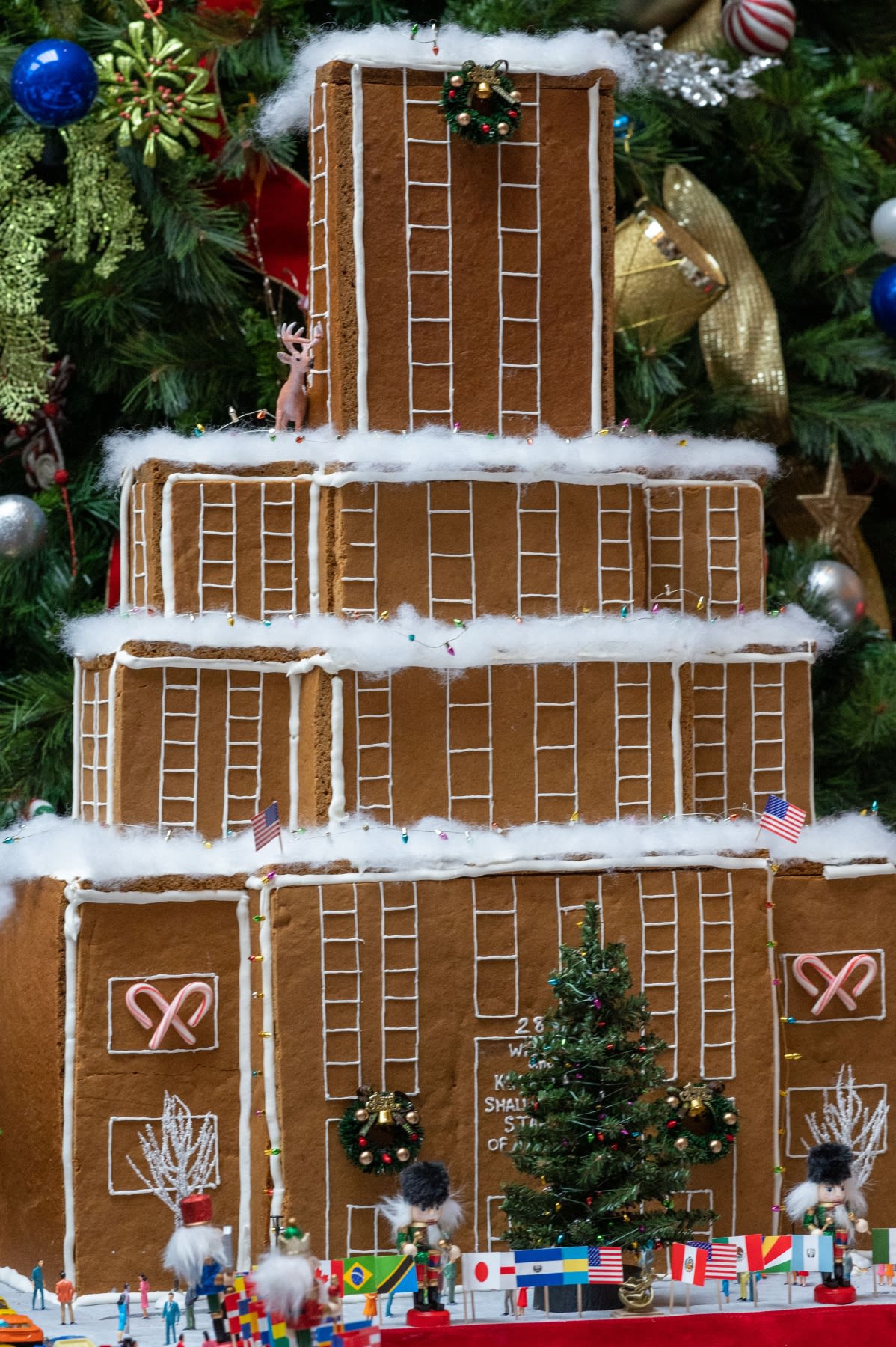 A three story gingerbread house with thing white lines of icing on the front decorated with miniature string lights and American flags.