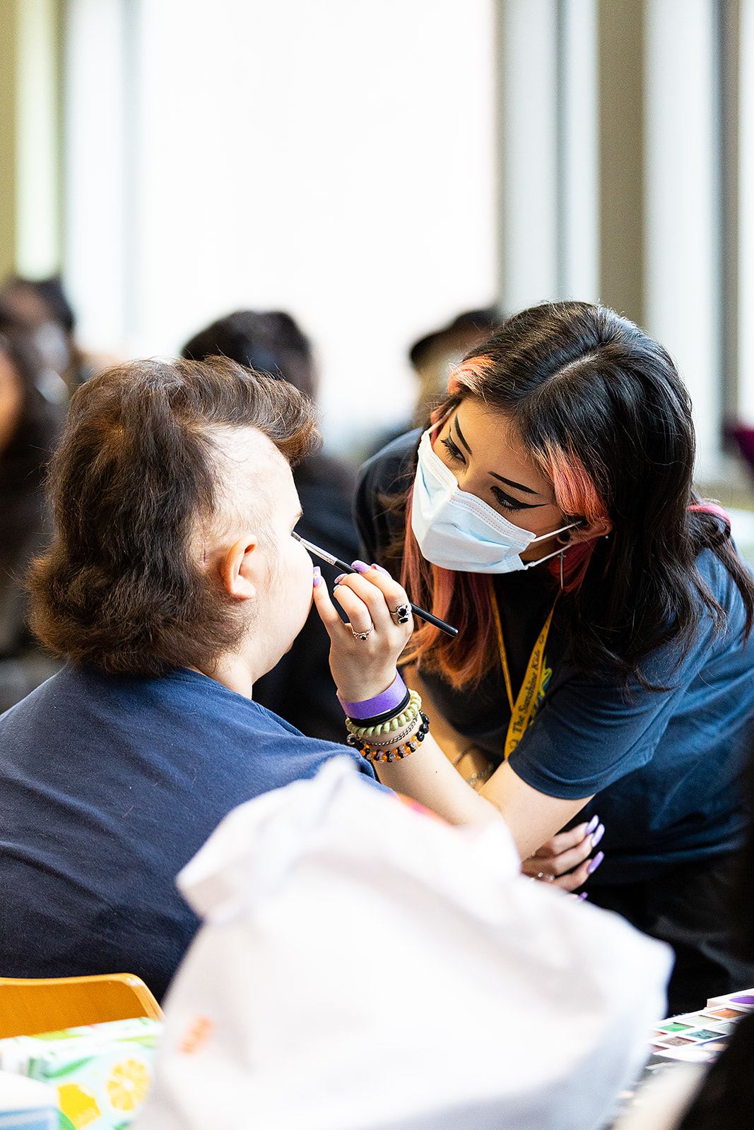 A person wearing a surgical face mask applying makeup to another person's face with a black makeup brush.