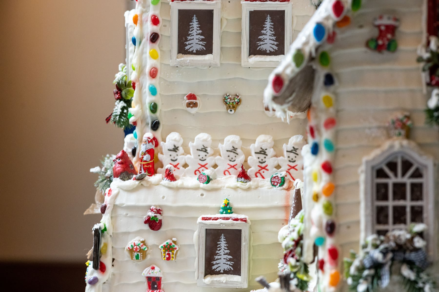 A gingerbread house that is completely coated in white frosting with little marshmallow candy snowmen on the side of it, along with jellybeans and other candies. 