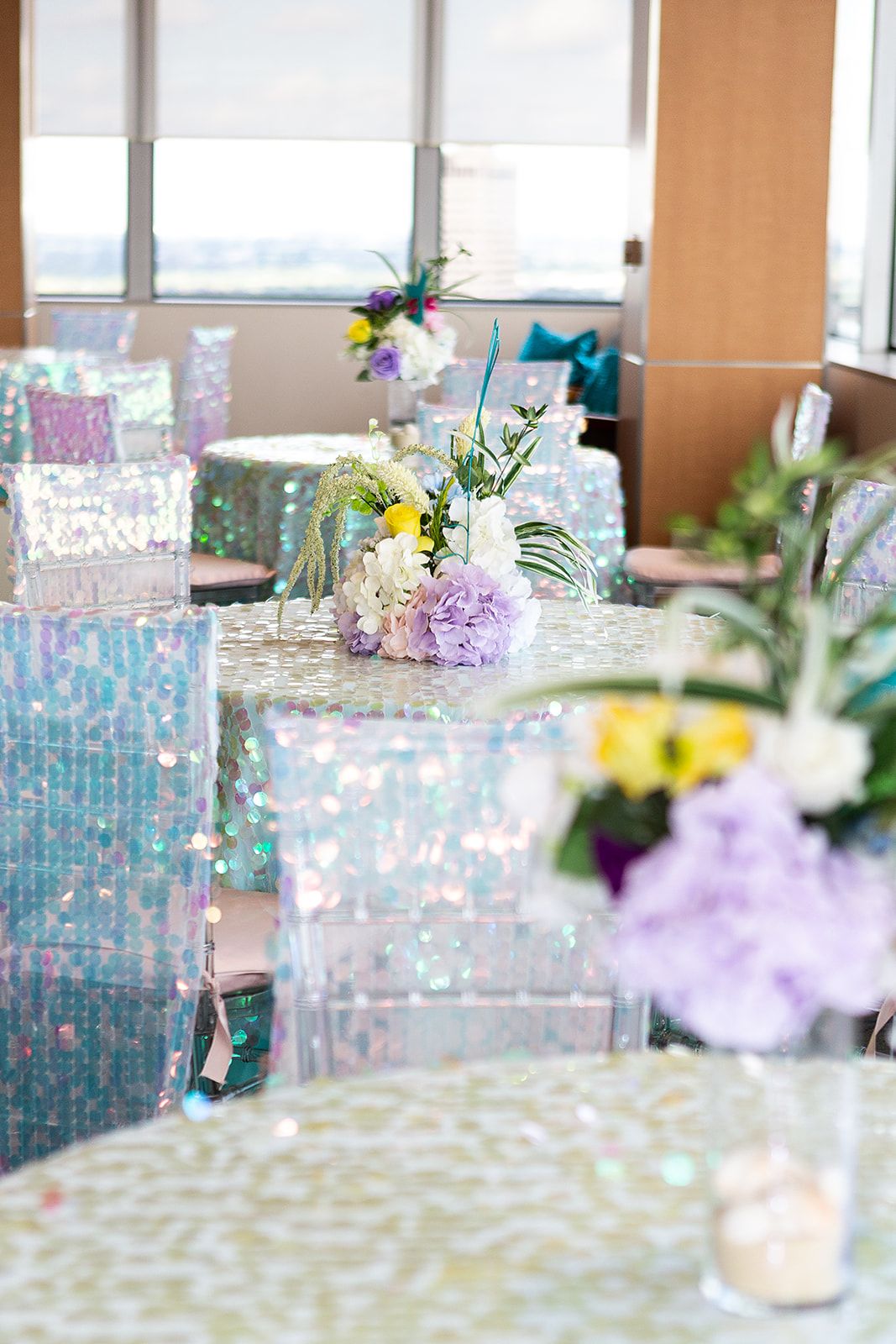 A purple. yellow, and white floral arrangement with light greenery on top of a blue and purple, iridescent sequin table cloth. 