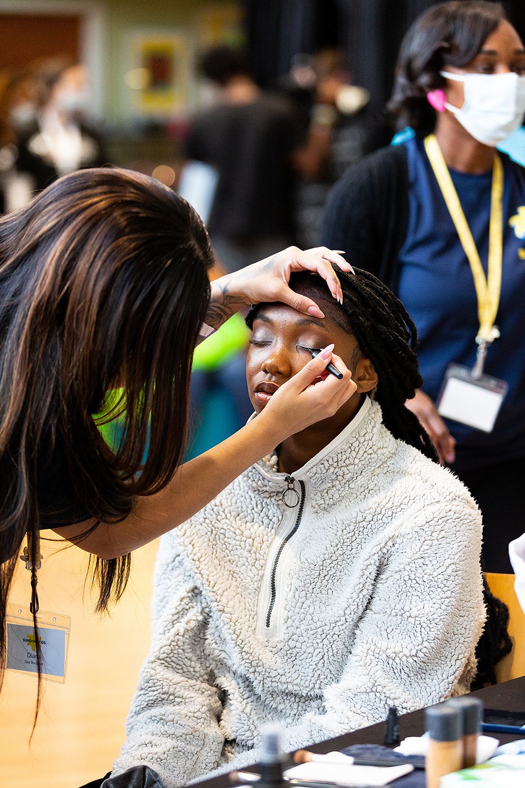  A prom attendee getting her makeup done. She is sitting with her eyes closed while another person is applying black liquid eyeliner to her top lash line.