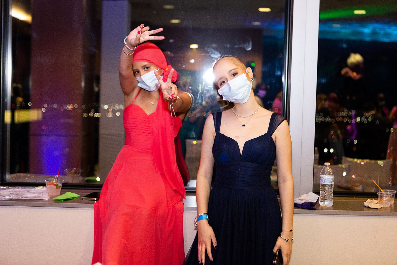 Two prom attendees posing for the camera. The girl on the left is wearing a red sleeveless gown with a matching headscarf. The girl on the left is wearing a navy sleeveless gown with layered necklaces.