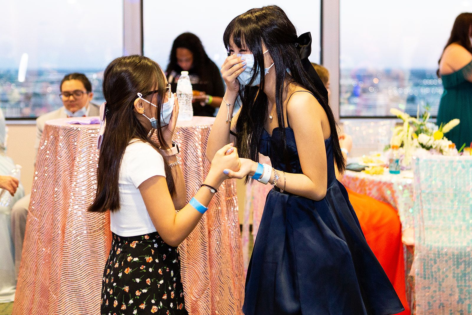 Two girls laughing together. One is wearing a white shirt with cap sleeves and glasses, and the other is wearing a navy blue strapless gown with a bow in her hair.