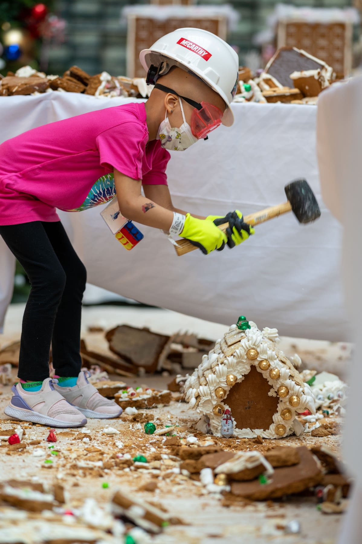 A person in a pink t-shirt wearing neon yellow gloves smashing a gingerbread house with a mallet.