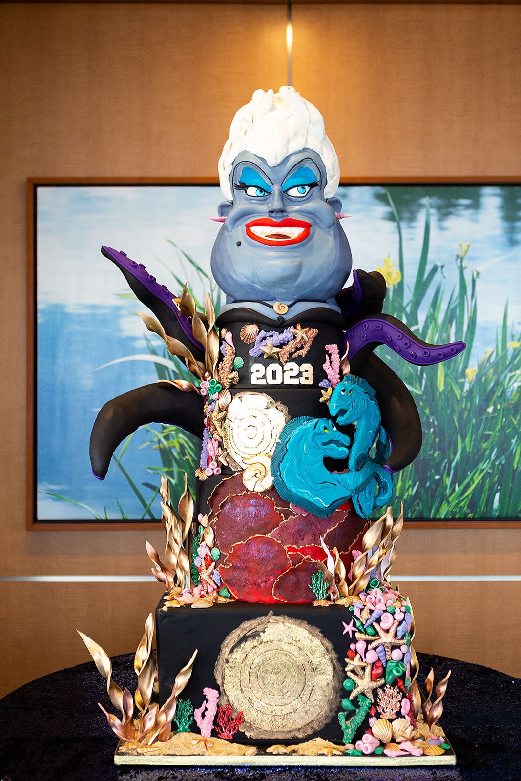 A two tier cake decorated in a Little Mermaid theme. The cake has Ursula's head on top with her tentacles coming out from the side. 2023 is on the front of the cake in white, surrounded by other aquatic decorations like sea shells, starfish, and corals.