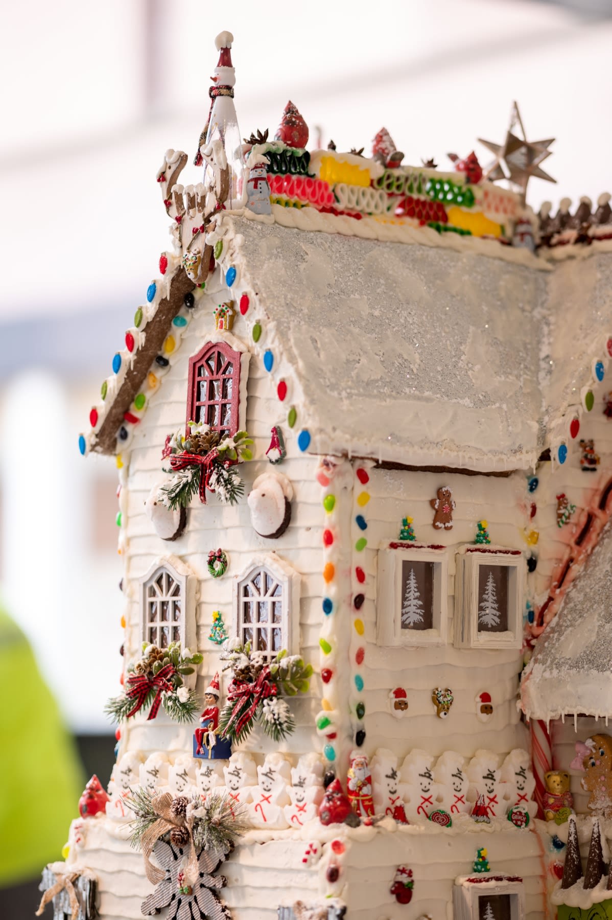 An all white gingerbread house with a red window and miniature green wreath on the front decorated with multicolor candies.