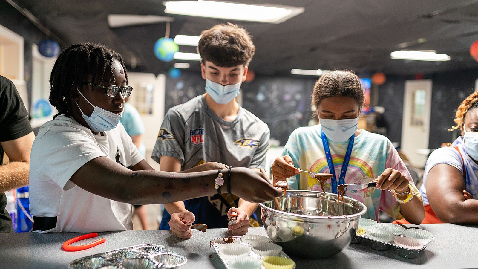 Three teens wearing face masks working together to scoop chocolate cake batter from a mixing bowl.
