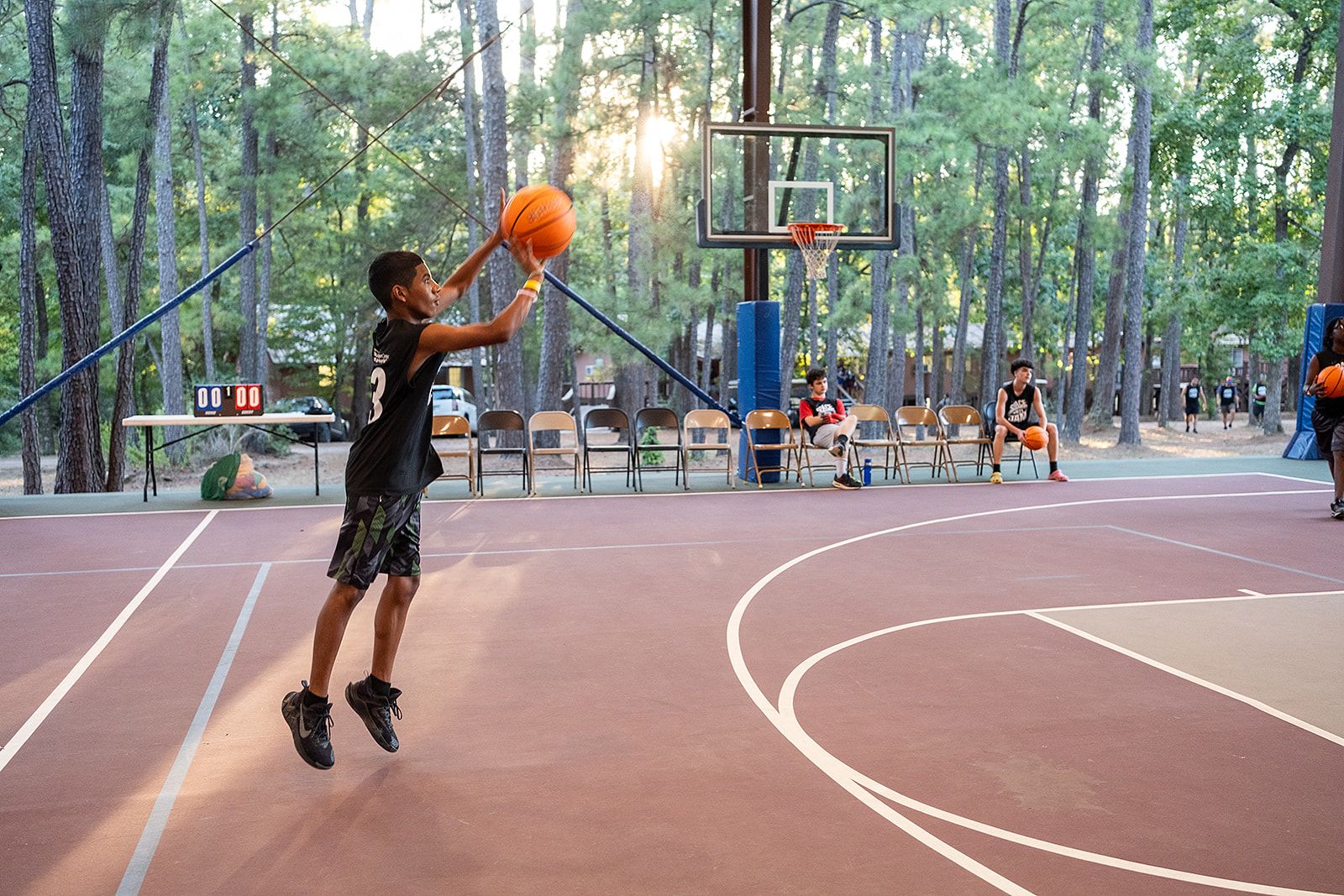 A kid wearing a black jersey shooting a basketball on a basketball court surrounded by trees.