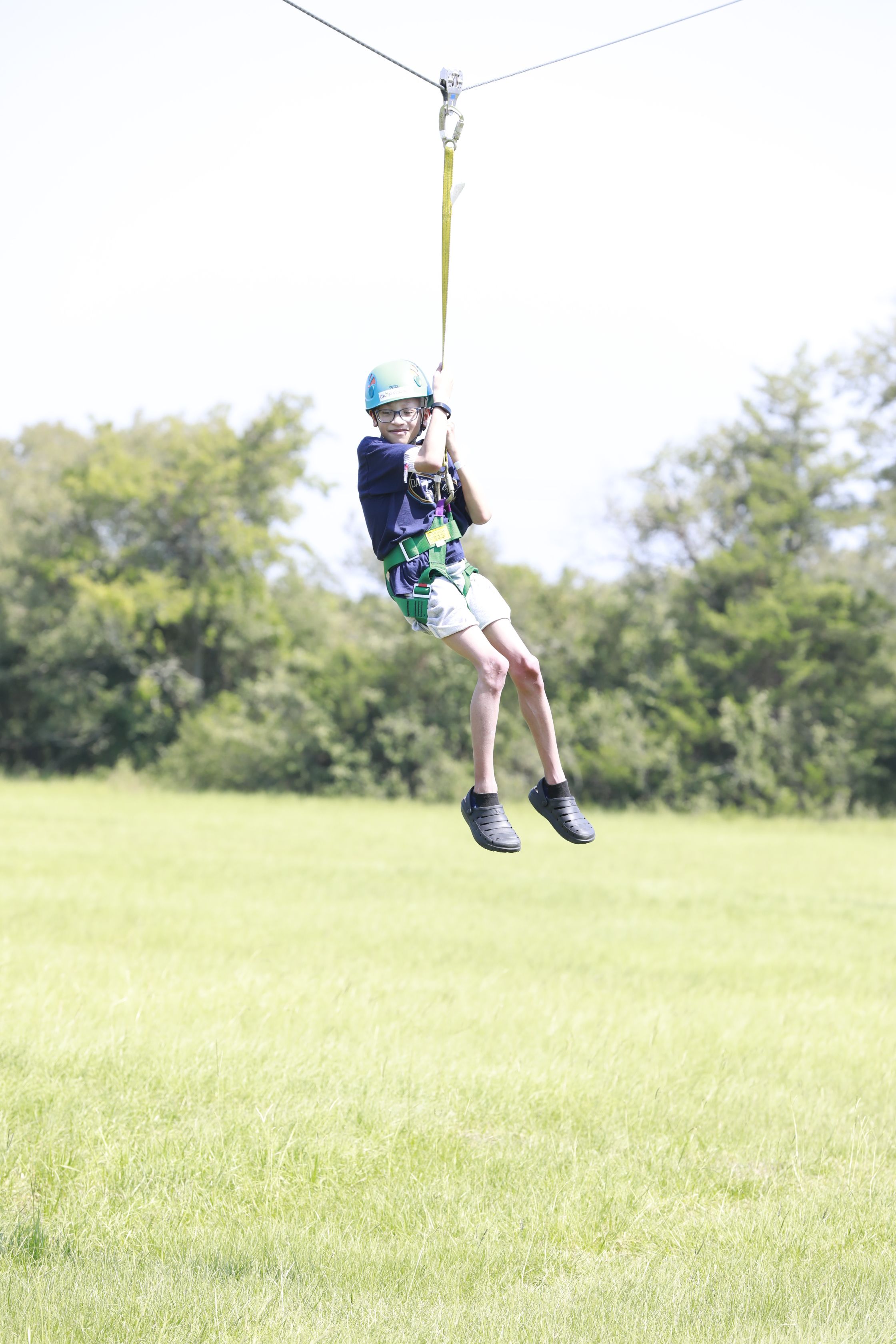 A child wearing a blue helmet is ziplining in a green harness.