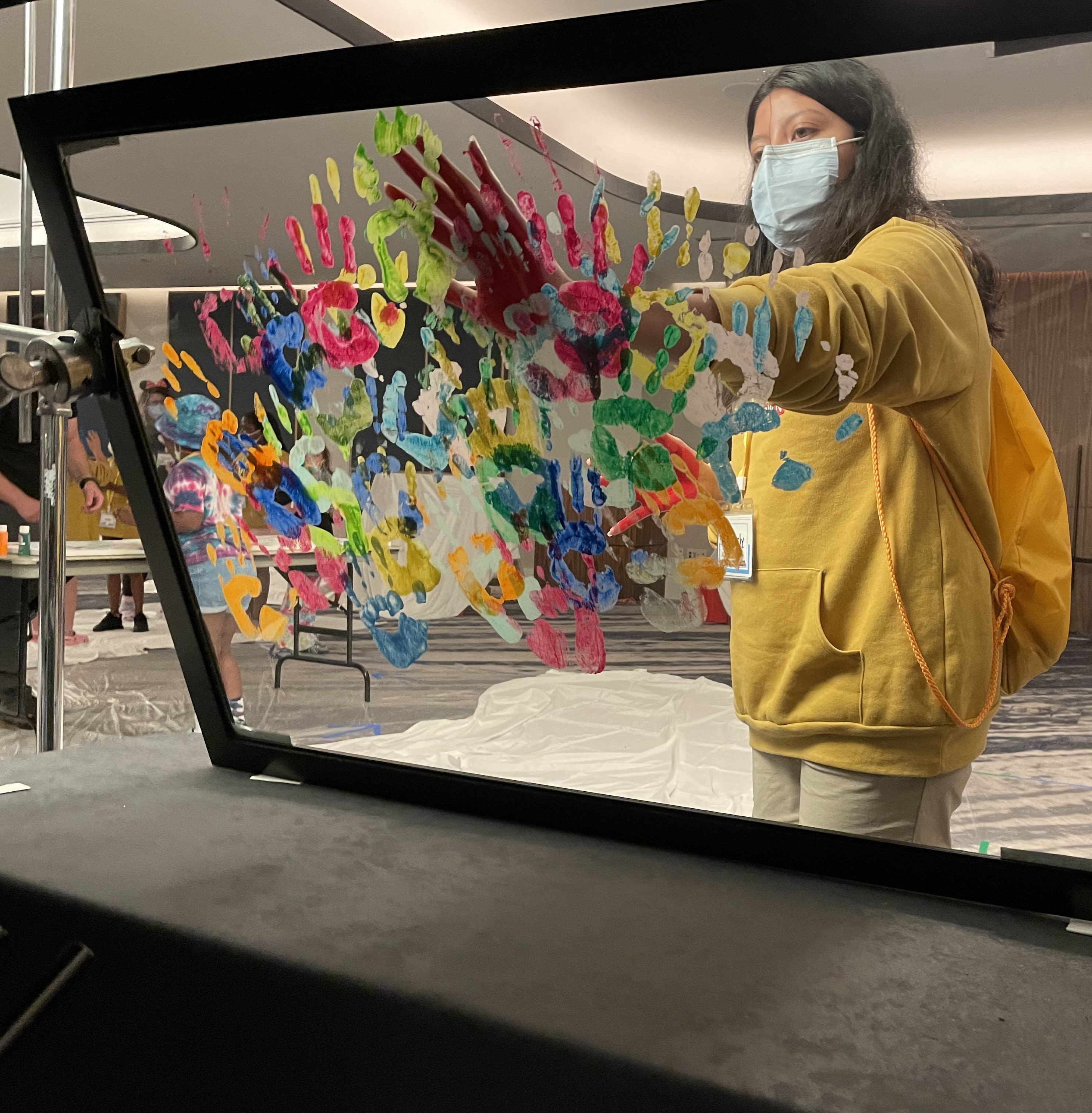 A child wearing a surgical face mask putting her handprint on a pane of glass that has many multicolored paint handprints on it.