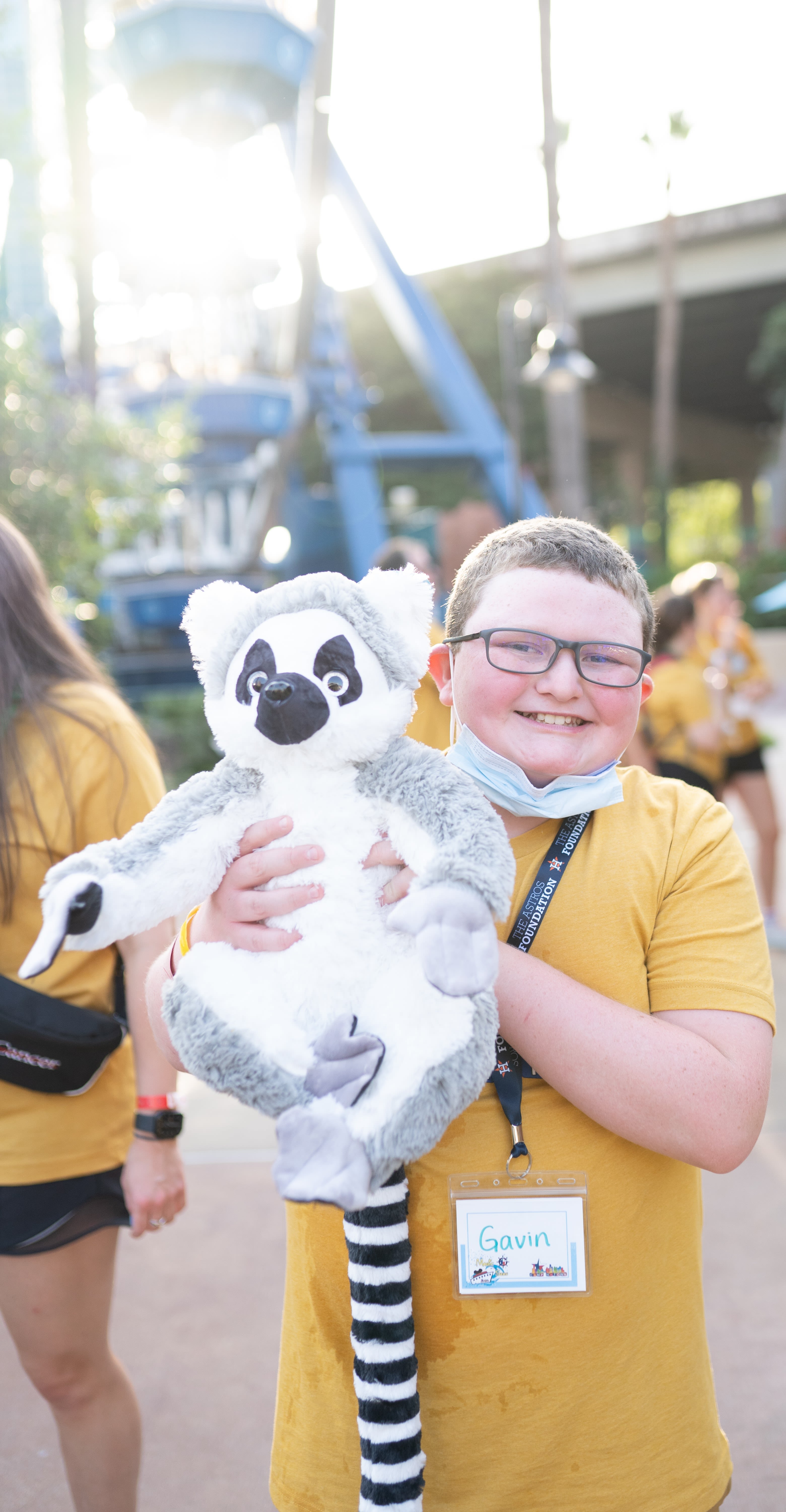 A child wearing a yellow shirt and glasses smiles while holding up a lemur stuffed animal. 