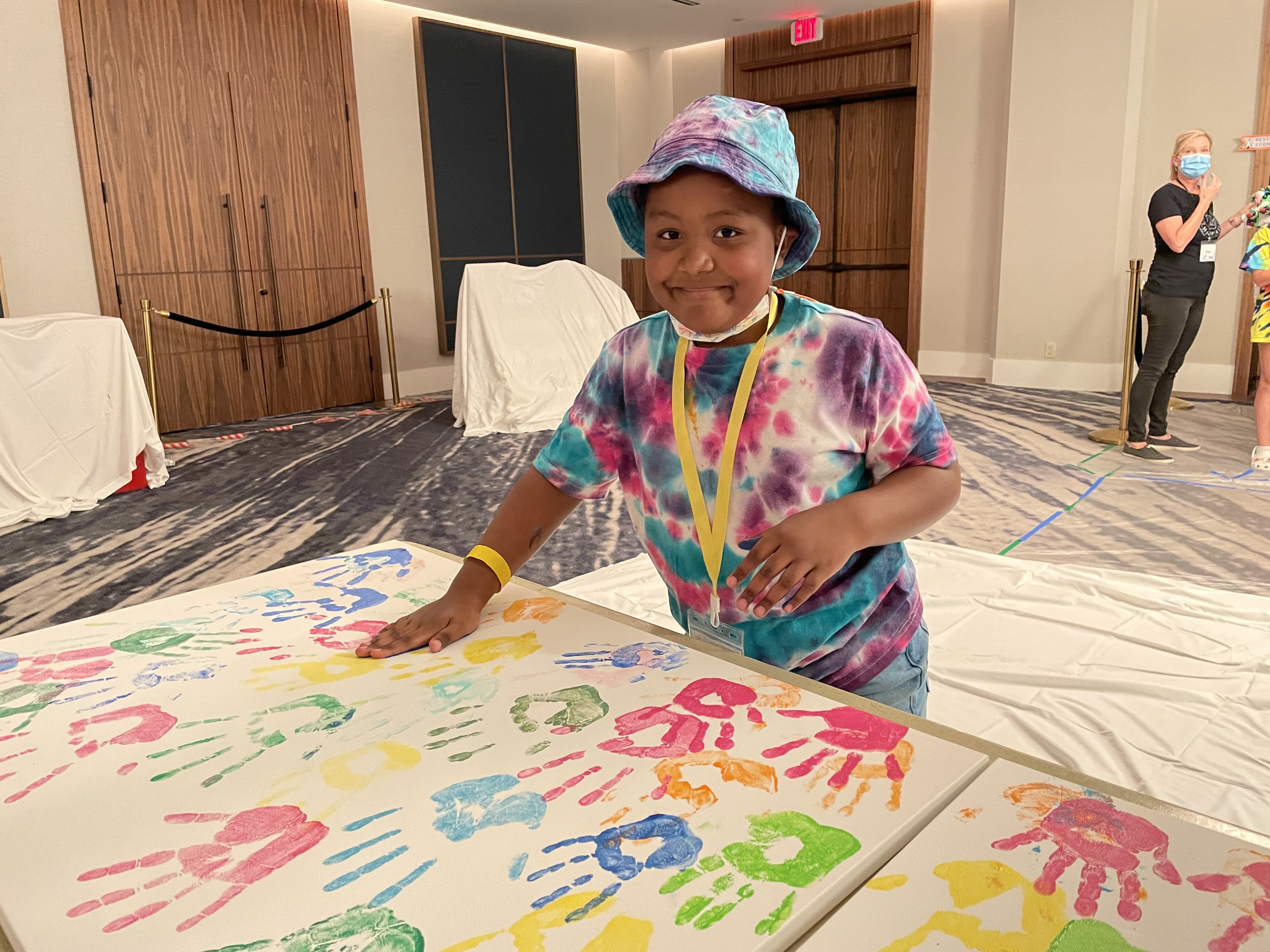 A child wearing a blue, pink, and purple tie dye t-shirt, smiling at the camera as they put their painted handprint onto a large white banner with other bright colored painted handprints. 