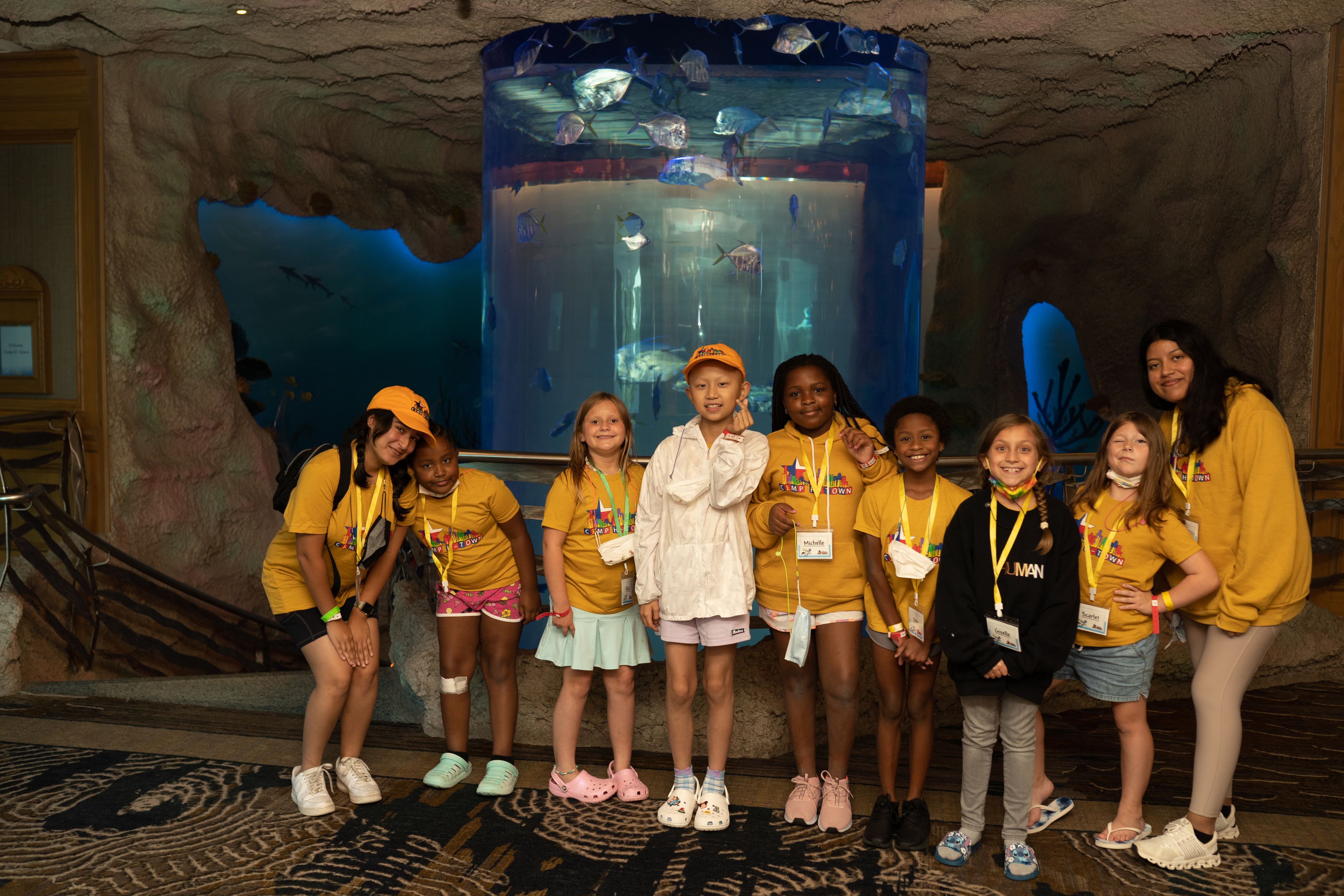 A group of kids in yellow shirts wearing yellow lanyards smiling in front of a large fish tank at an aquarium.