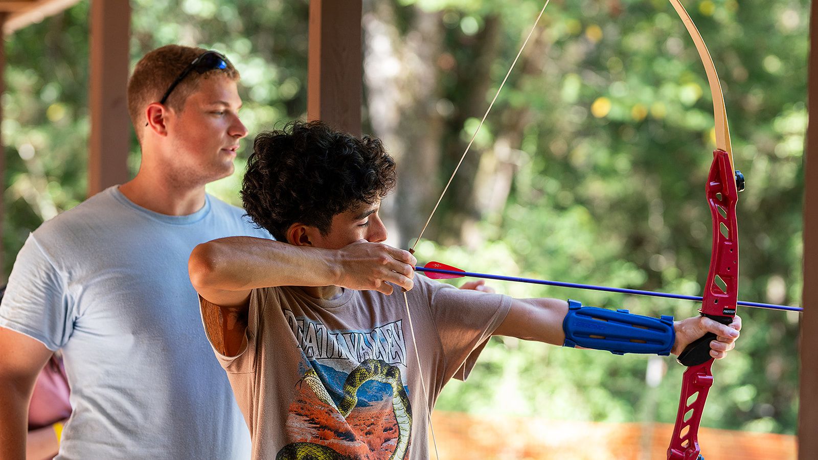 A person pulling the arrow back on an archery bow, with another person standing behind them.