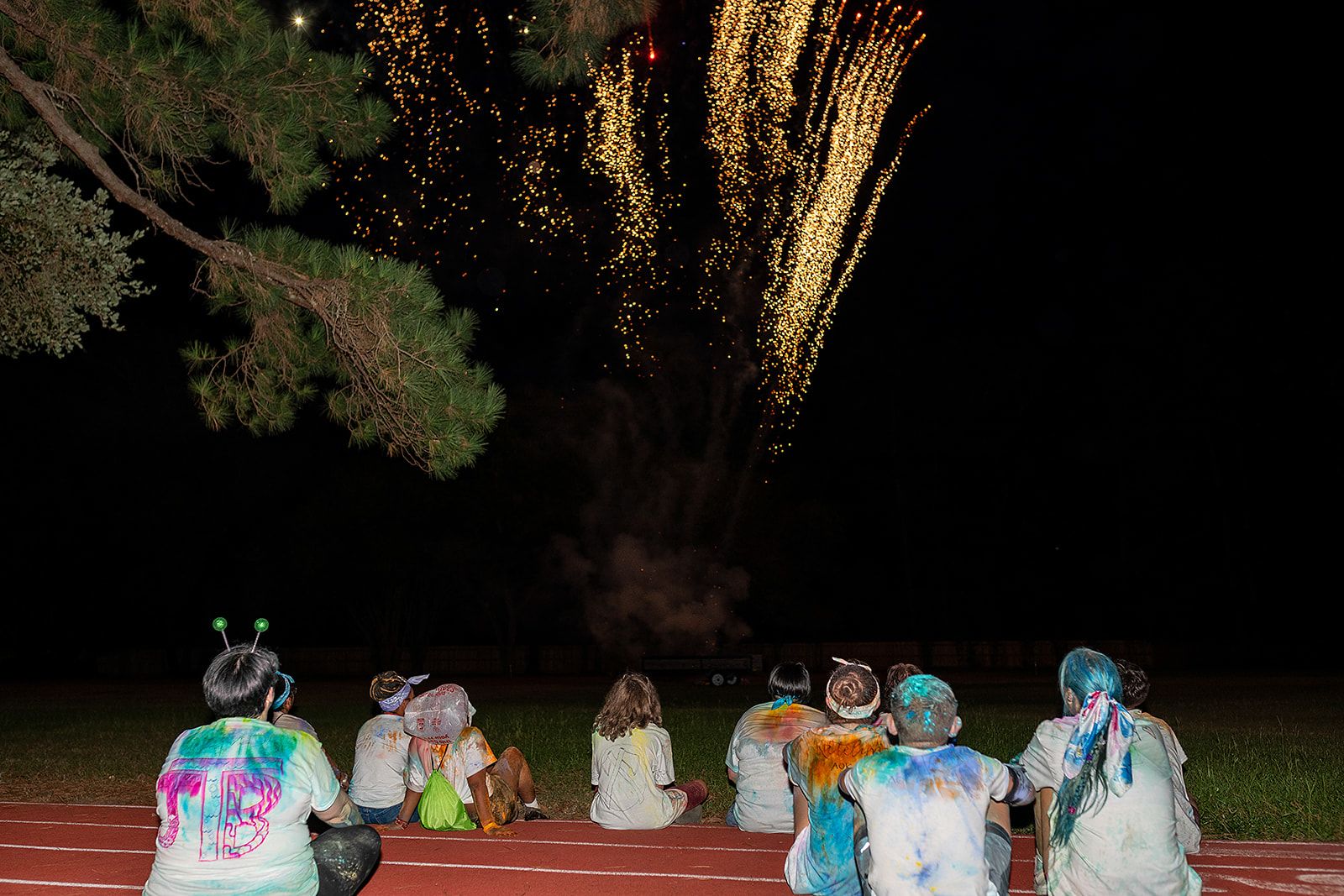 Several people wearing white shirts covered in multicolor powder sitting on the ground watching fireworks.