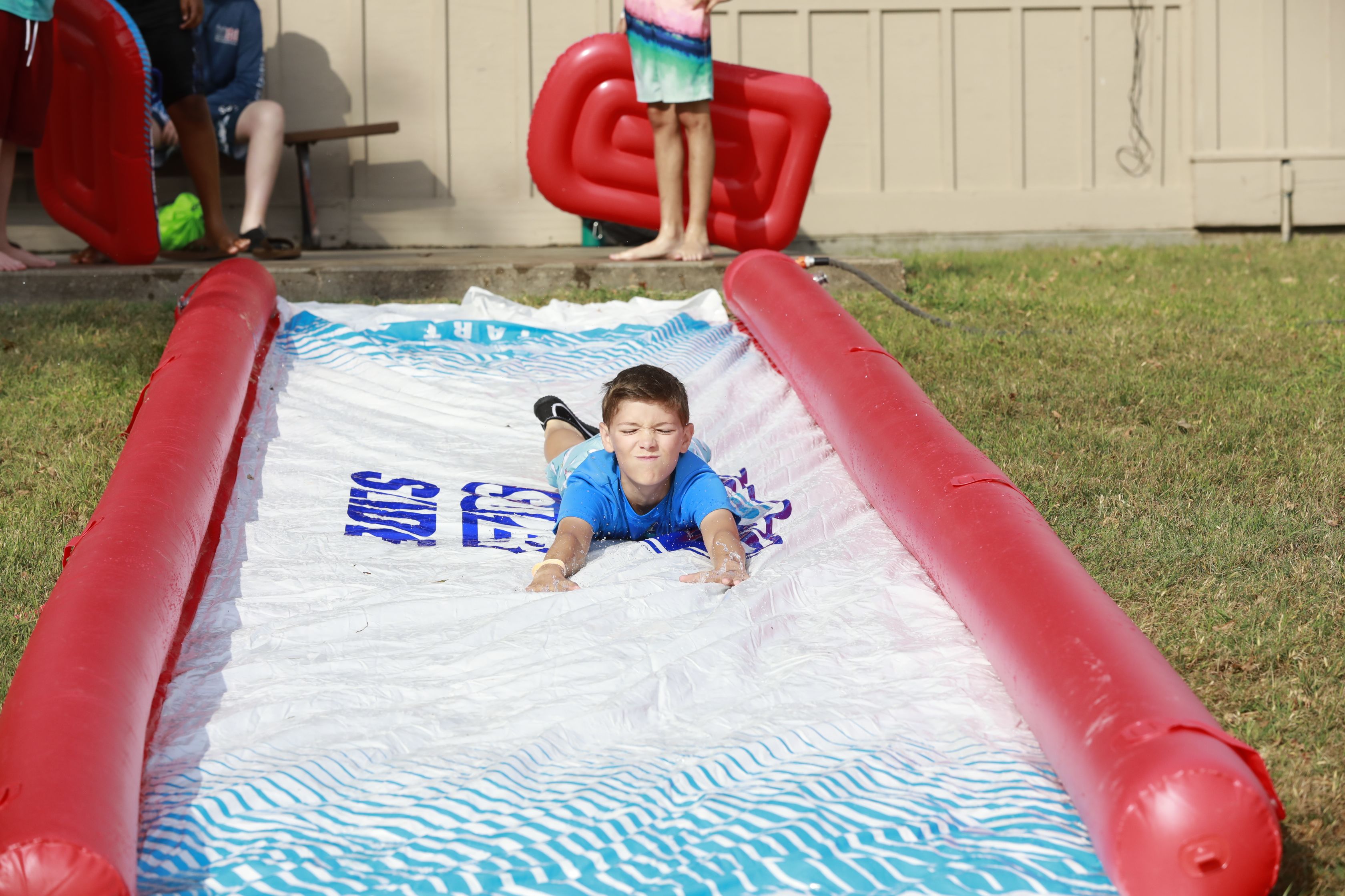 A boy wearing a blue shirt and swim trunks sliding with his arms out down a slip and slide.