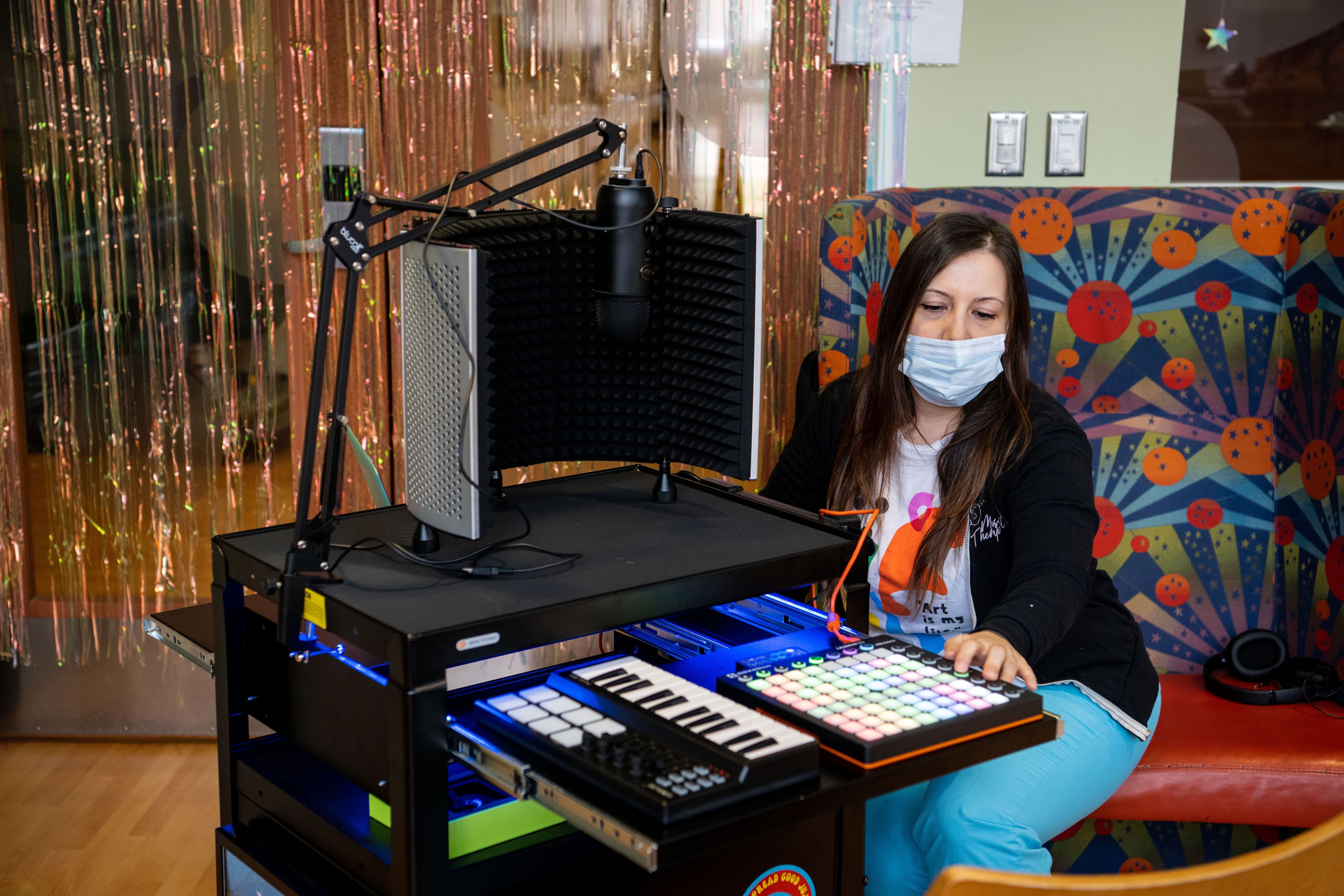 A person wearing a surgical face mask pushing a button on a soundboard, sitting at a table with a small keyboard and microphone on it.