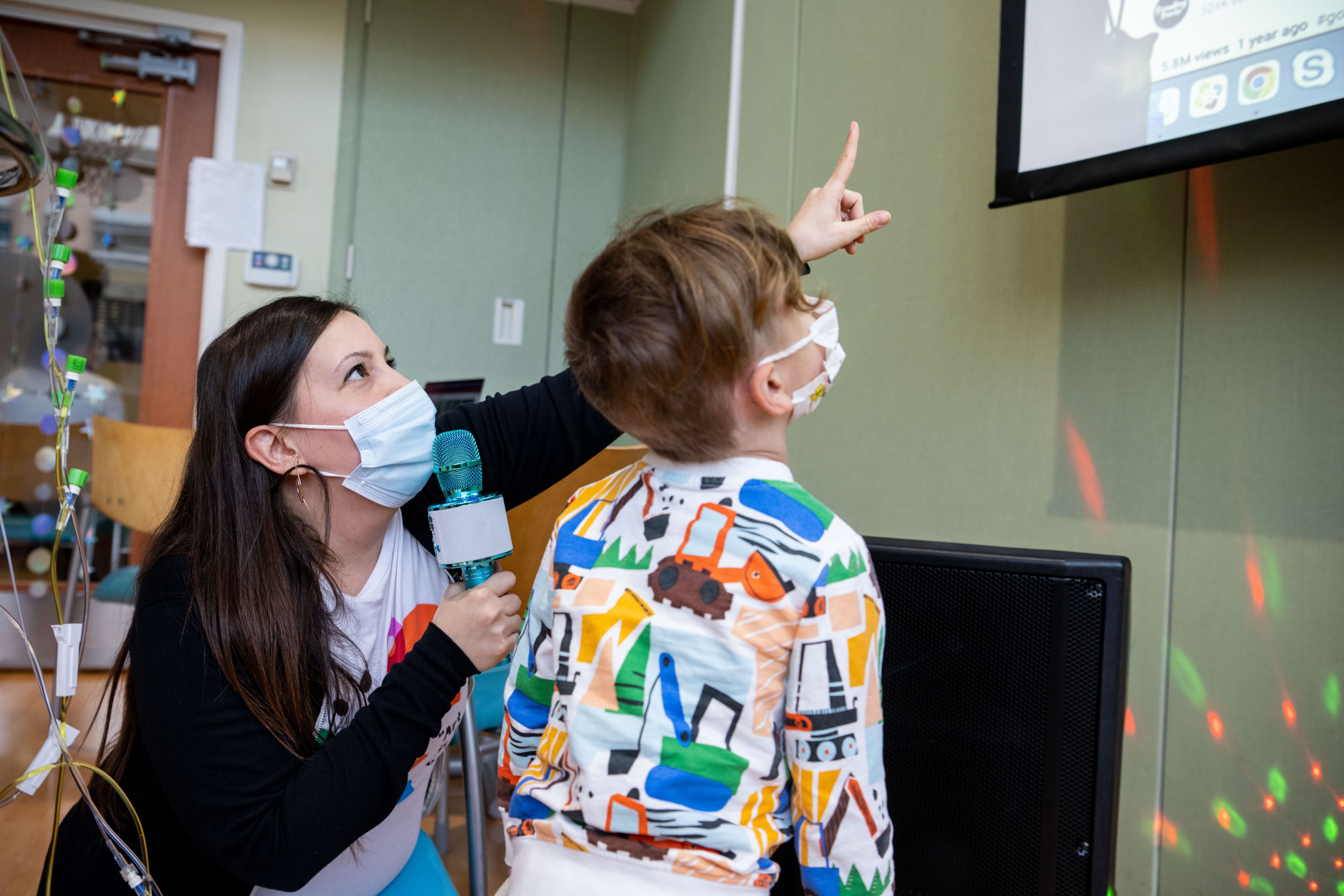 Young boy in colorful pajamas and a mask looking up at a screen, while a woman with a microphone and mask kneels beside him pointing at the screen.