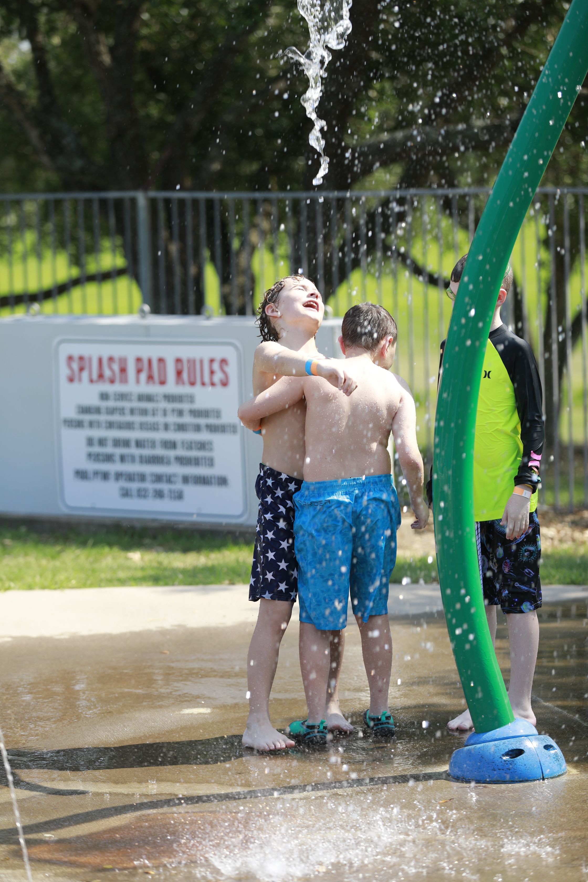 Two boys with their arm around each other standing under falling water at a splash pad.
