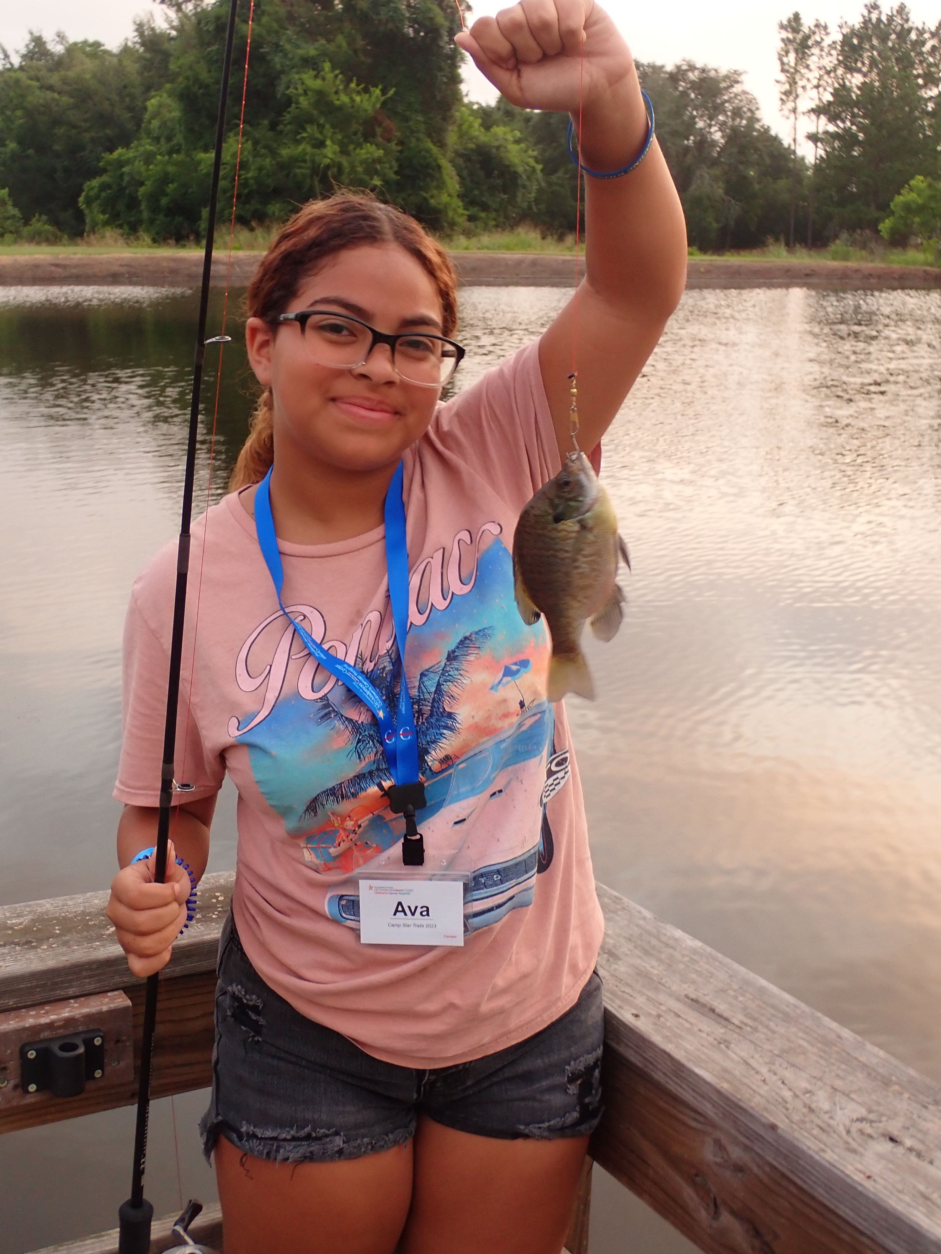 A girl wearing black glasses holds up a fish she caught on a fishing line in front of a lake.