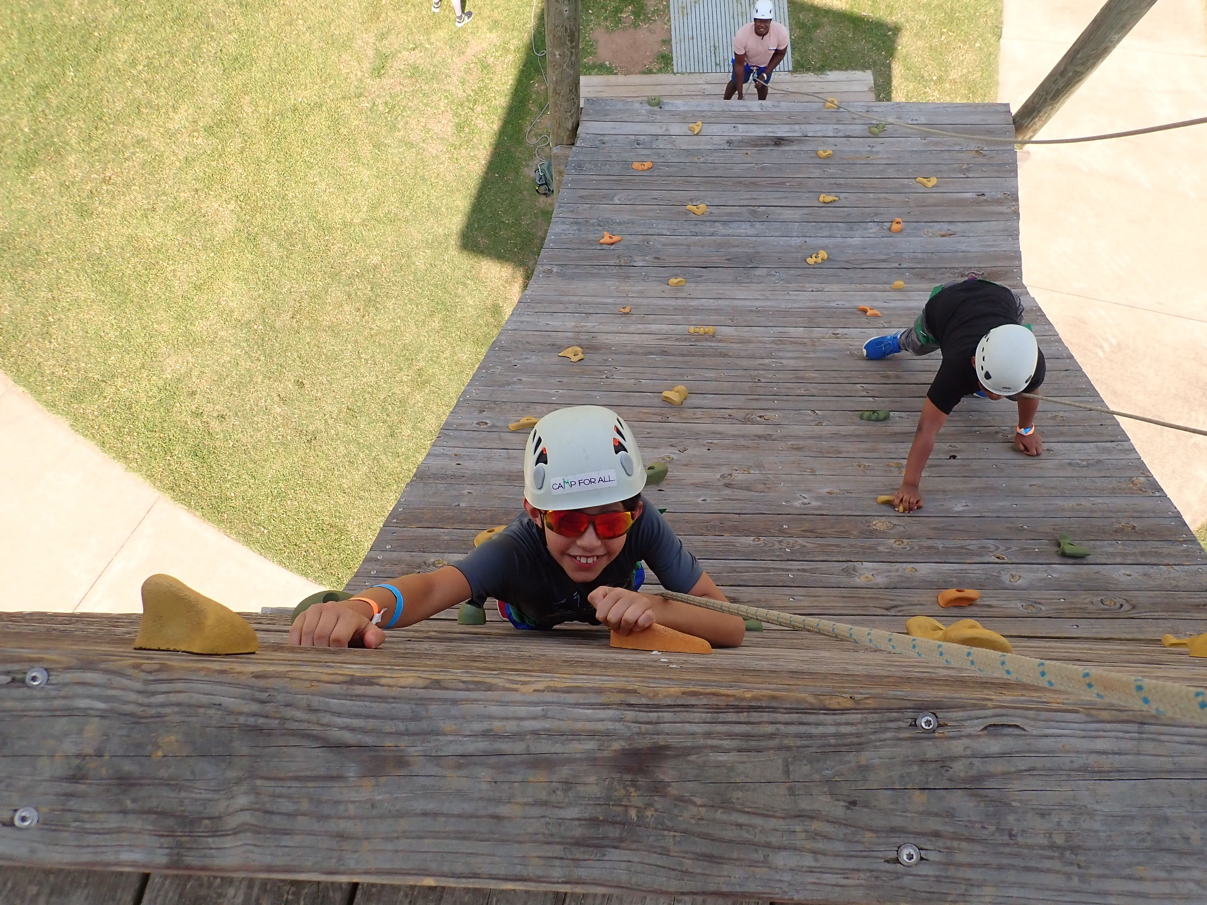 A child wearing a helmet and sunglasses looking up while on a climbing wall.