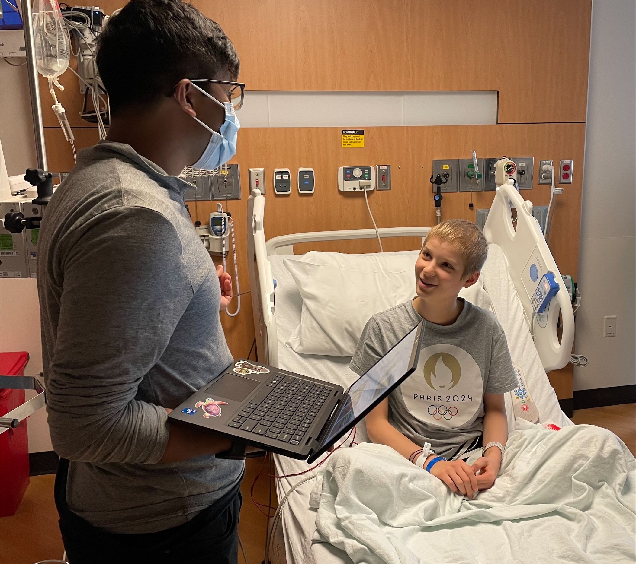 A child sitting up in a hospital bed talking to a hospital staff member, who is holding a laptop.