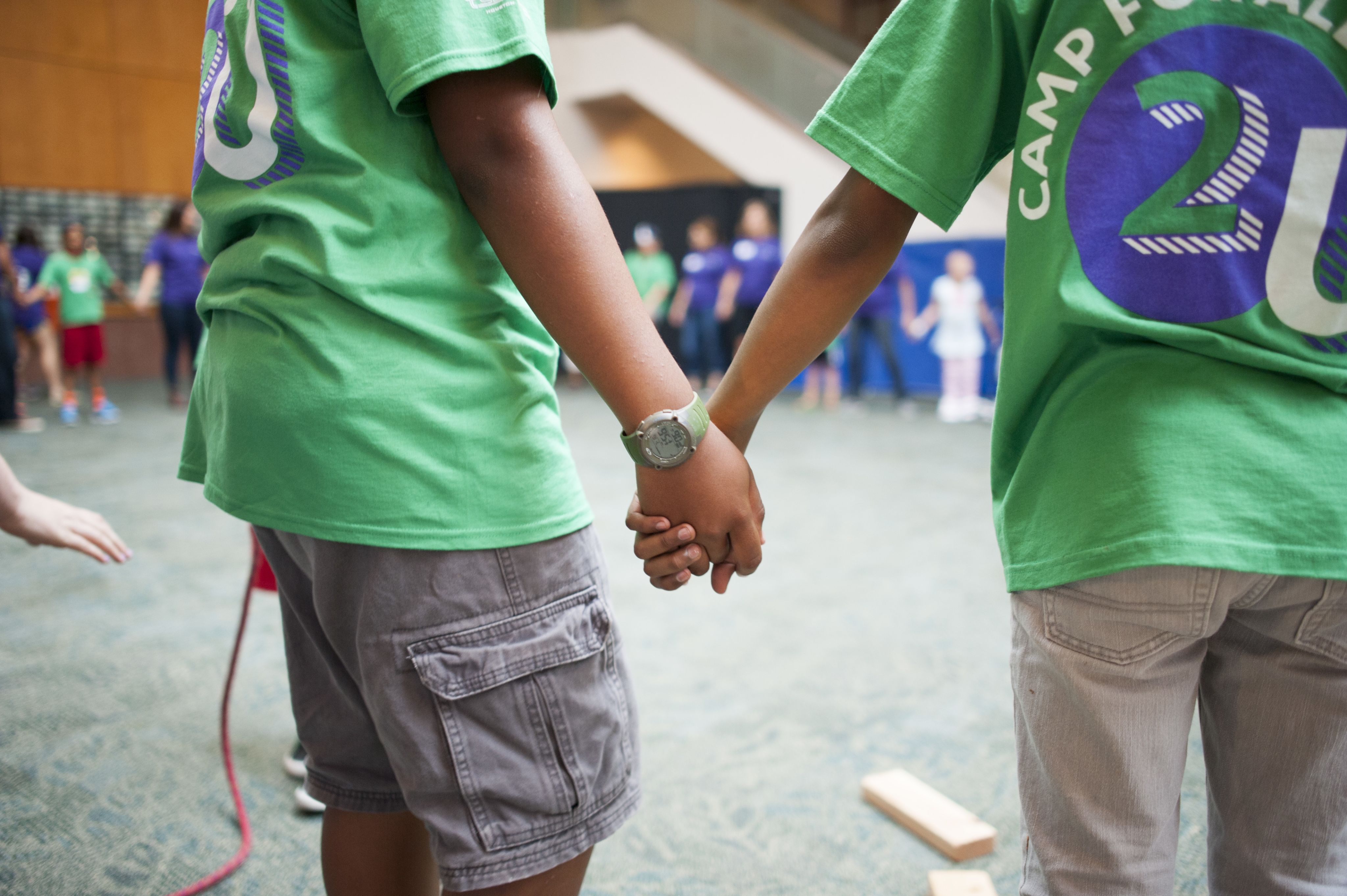 Two people in green t-shirts holding hands in a large room with other people in the background.
