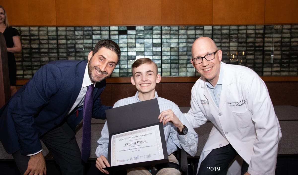 Three people posing together at an award ceremony. A young man in the center is holding a certificate of scholarship and smiling, with a man in a suit on the left and a man in a white lab coat on the right.