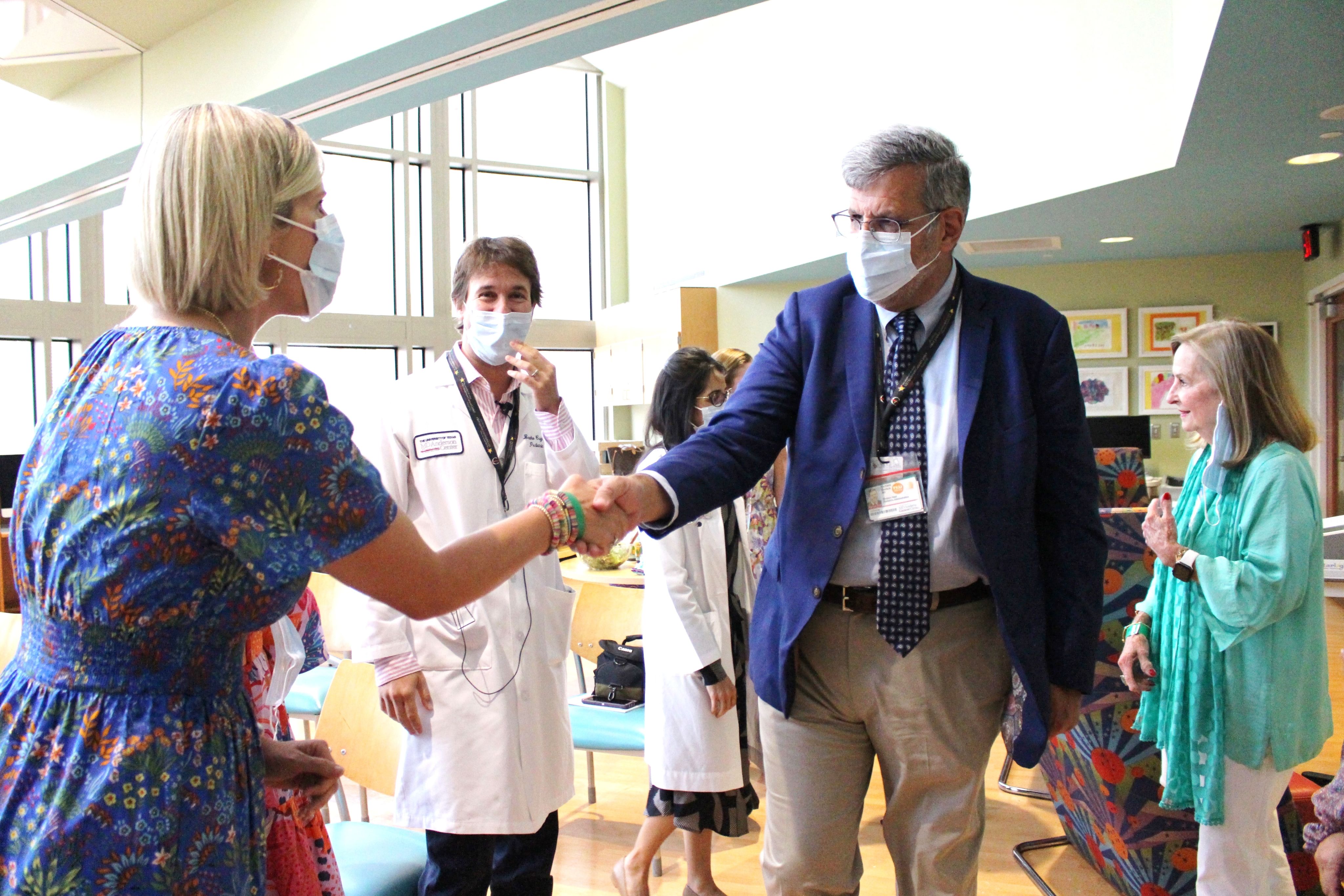 Dr. Richard Gorlick in a suit and mask shaking hands with a woman in a colorful dress and mask, with medical staff and others in the background.